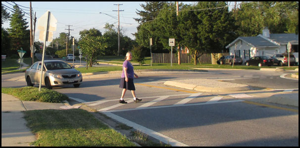Photo shows a woman walking across the first of two lanes with an island between the lanes.  About 10 feet to our left, her lane exits a roundabout (lanes go around a large circle).  A car is coming from the roundabout and is waiting for her while she looks to her right for cars coming in the next lane.