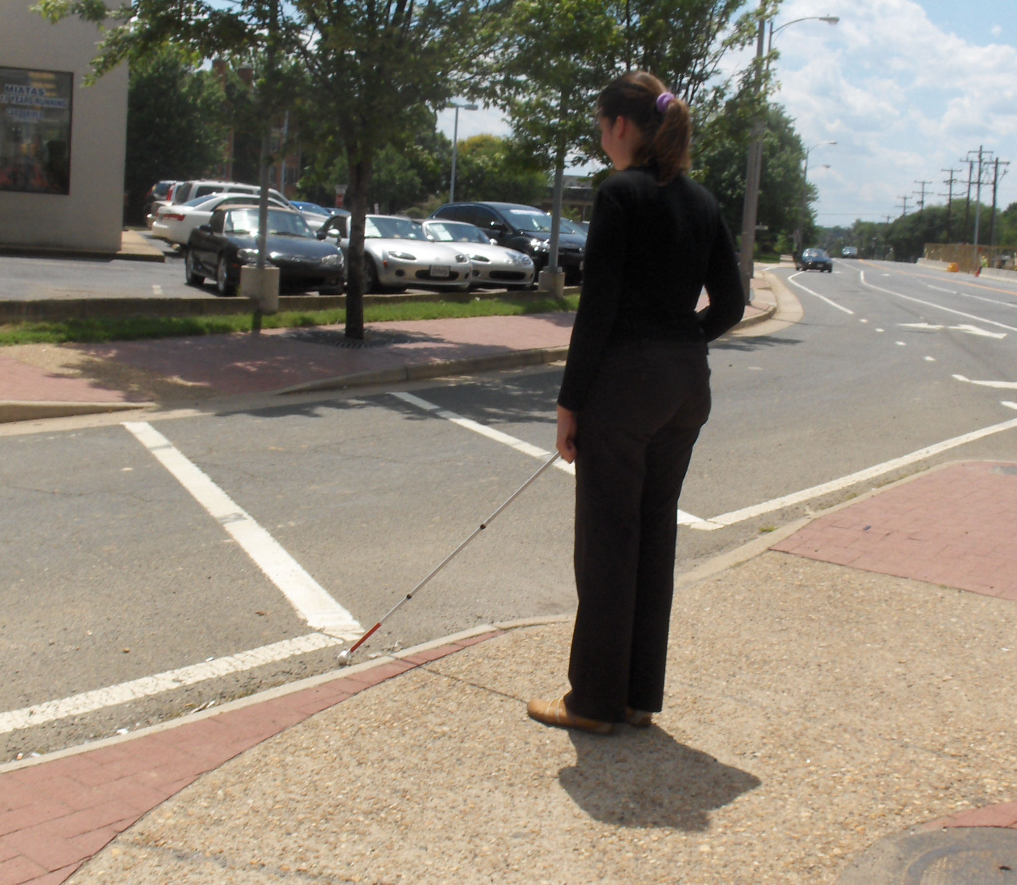 Two photos show Mimi standing on the SW island with her cane near the crosswalk of the right-turning lane. To her right is a street with one or two cars.  The second photo shows a minivan leaving the street and turning slightly to its right to enter the lane in front of Mimi and approach the crosswalk.