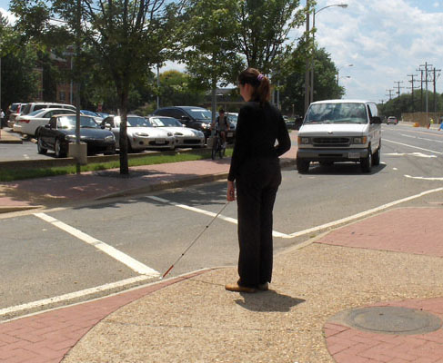Two photos show Mimi standing on the SW island with her cane near the crosswalk of the right-turning lane.  To her right is a street with one or two cars.  The second photo shows a minivan leaving the street and turning slightly to its right to enter the lane in front of Mimi and approach the crosswalk.