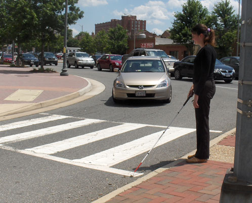Two photos show Mimi standing on the SE island with her cane in the crosswalk of the right-turning lane.  To her right is a very busy street.  The second photo shows a car turning right from the busy street almost 90 degrees into the lane in front of Mimi and approaching the crosswalk.