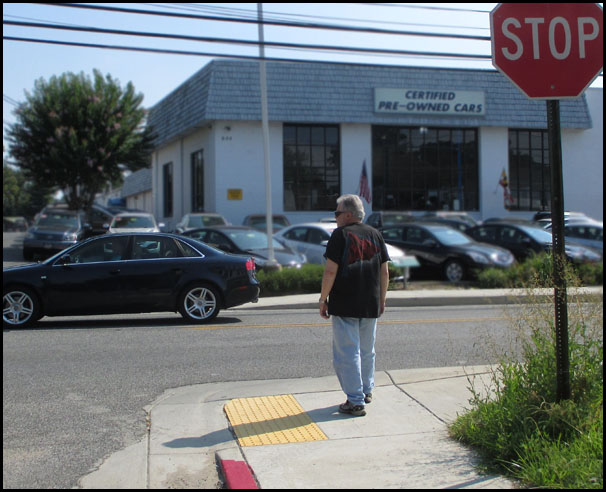 Photo shows a man starting to walk across a two-lane street to a used car dealer.  We can see the stop sign for the street beside him.