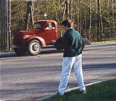 photo shows the boy with a white cane in one hand and a stopwatch in the other.  He is standing at the curb of a street that has a bend to the right.  He is looking at a truck that has just come around the bend on the right.