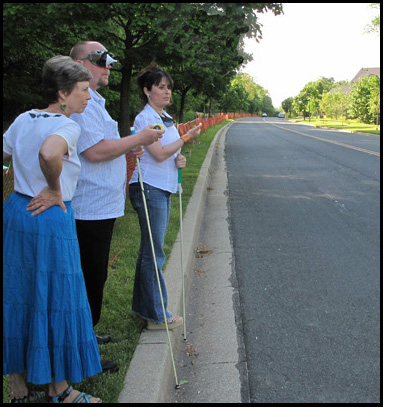 3 photos show Paul and Jomania standing at the curb of a straight street, holding stopwatches -- Paul is wearing a vision simulator for restricted visual fields.  Dona stands to their right.  They are both watching a car approach from the left and pass them.