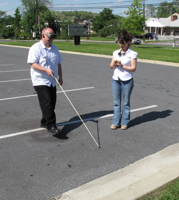 Photo shows Paul wearing opaque goggles and walking with a cane across the lanes in the parking lot, while Jomania looks at a stopwatch.
