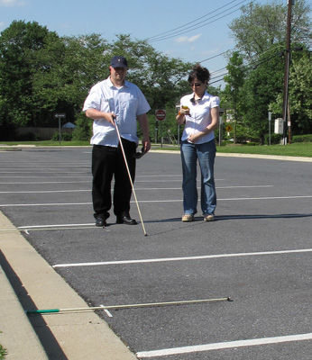 Photo shows instructor and student standing and talking at the far side of the second lane in the parking lot, the instructor is looking at a stopwatch.