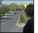 Photo shows a woman standing on a median of a 6-lane street and looking to her right at traffic approaching about a block and a half away.