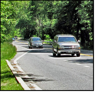 Photo shows a two-lane road that appears from around a bend about a block away -- it widens to 3 lanes just before it reaches us.  A long string of cars approaches us but some of them are obscured on the left by trees and bushes that overhang the street.