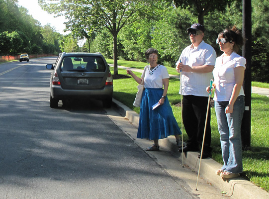 Photo shows Paul and Jomania standing at the curb of a wide two-lane street with our vehicle parked to their right. Dona is pointing to a car coming from their right.