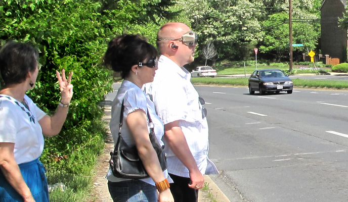 2 photos show two students wearing occluders at the edge of a two-way street with 4 lanes coming from the left, 3 lanes from the right.  Each photo has a car in one of the lanes from the left. Dona is behind the students and holding up fingers indicating whether the car is in the first, second, or third lane.