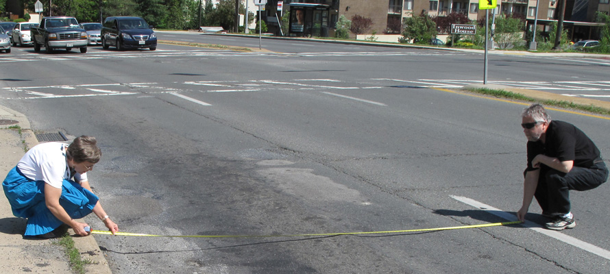 Dona stoops at the curb of a 6-lane highway holding one end of a tape measure while Fred holds the other end at the white line marking the first lane.