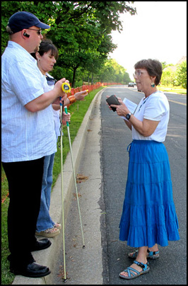 Photo shows Dona holding a small tape recorder and standing in a residential street talking to two people who are standing on the curb.