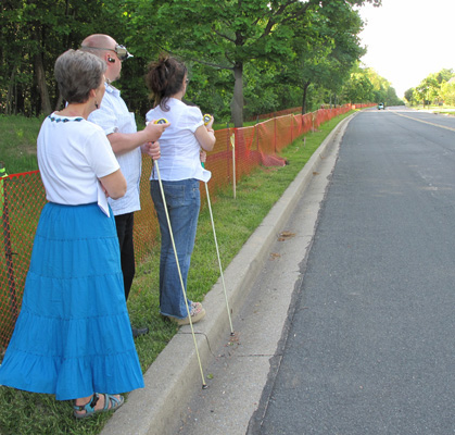 3 photos show Paul and Jomania standing at the curb of a straight street, holding stopwatches -- Paul is wearing a vision simulator for restricted visual fields.  Dona stands to their right.  First they face forward, then they turn to the left and see a car approaching from a distance, then turn back and face forward again.