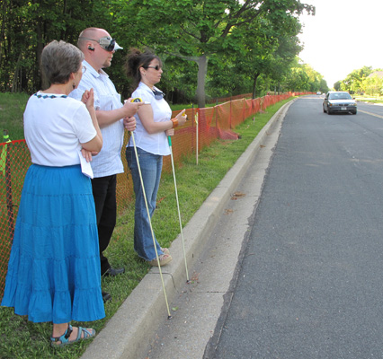 3 photos show Paul and Jomania standing at the curb of a straight street, holding stopwatches -- Paul is wearing a vision simulator for restricted visual fields.  Dona stands to their right.  First they face forward, then they turn to the left and see a car approaching from a distance, then turn back and face forward again.