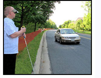 Photo shows a man wearing a funnel-shaped visual simulator, standing at a curb, holding a cane and wearing a vision simulator for narrow visual field.  In the street, about 20-30 feet to his left is a car approaching him.