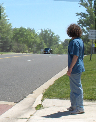 Photo shows a student at a crosswalk, looking to the right.  A dark truck appears at the top of the hill on the right.