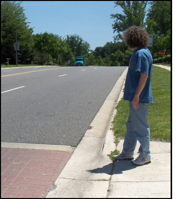 Photo shows the street to the right of the crosswalk, gently sloping up to peak about 3/4 of a block away, and a blue truck appears at the top.  Stephan, a young adult, is standing at the edge of the crosswalk and looking to the right.