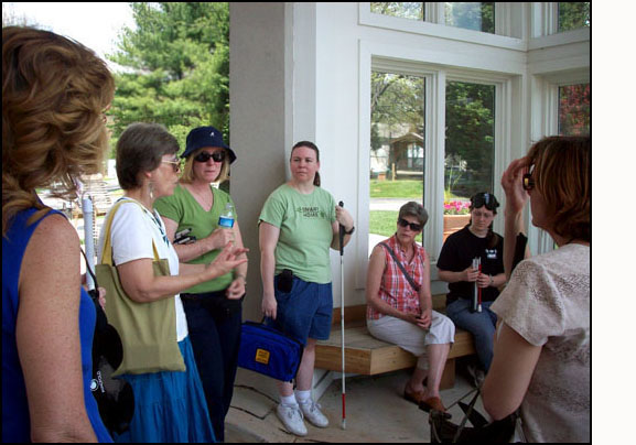 Photo shows Dona talking with the participants, we can see 6 of them, each is holding a white cane and one has a blindfold on the top of her head. They are standing and sitting in a gazebo with a parking lot behind them.